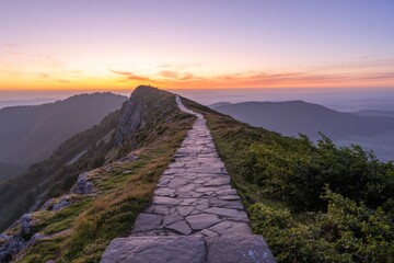 A narrow, winding stone path leads along a majestic mountain ridge, illuminated by the warm glow of a sunset. 