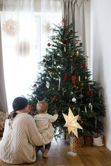 Stylish mother and cute baby sitting at illuminated star and decorated christmas tree with lights....