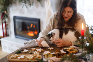 Woman together with cute cat decorating christmas gingerbread cookies with sugar glaze on rustic...