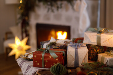 Merry Christmas! Stylish wrapped christmas presents, ornaments, candle, gingerbread cookies on table against festive decorated fireplace with lights. Atmospheric holiday eve