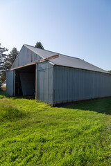 Gray Metal Pole Barn Farm Shed With Sliding Door Open Surrounded by Green Grass with Evergreen...