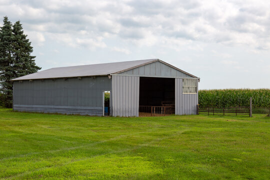 Gray Metal Pole Barn Farm Shed With Sliding Door Open Surrounded by Green Mowed Grass and a Tasseled Cornfield