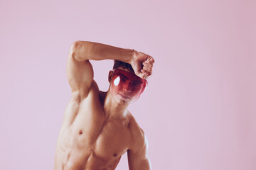 Confident young man posing with sunglasses, showcasing a fit physique against a pastel pink...
