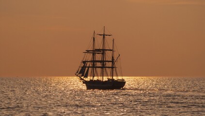 Obraz premium Gliding three-masted ship on calm open ocean under orange sunset sky, rigging and spars sparkling
