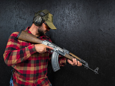 A man in a plaid shirt wearing safety gear inspects a classic russian old assault rifle.