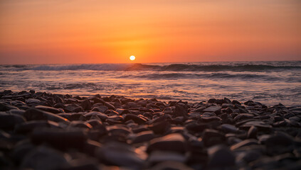 The sun sets over the ocean, casting an orange glow on the waves and rocks.