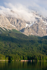 Eibsee Lake in Bavaria, Germany, with small lakeside houses and cabins at the forest edge, beneath the towering Zugspitze mountains, partly covered by clouds. A scenic alpine landscape.