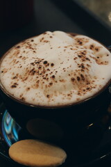 Close up of a hot cappuccino coffee in a dark mug. Frothy milk foam with chocolate powder and a...