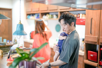 In a Mediterranean-style dining kitchen, a Japanese man in his 30s is holding a three-month-old baby while wiping the table with a cloth. Summer. Japan.