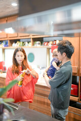 In a Mediterranean-style dining kitchen, a Japanese father in his 30s holds a three-month-old baby while wiping the table and talking to the mother in her 20s wearing orange clothes. Summer. Japan.