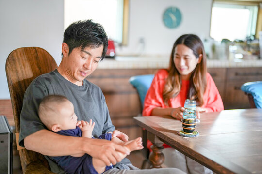 A Japanese father in his 30s holds a three-month-old baby while sitting at a Mediterranean-style dining table, sharing coffee and conversation with the mother in her 20s. Summer. Japan.