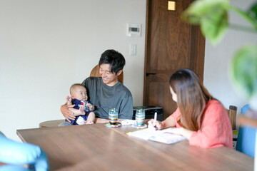 Fototapeta premium A man and a woman are sitting at a table with a baby in front of them. The man is holding the baby while the woman is writing something. Scene is warm and loving