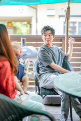 A man sits at a table with two other people, one of whom is holding a baby. The man is wearing a...