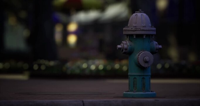 A blue hydrant is placed prominently on the street, surrounded by blurred urban greenery. Soft light from nearby sources creates a peaceful nighttime atmosphere.