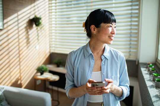 Asian woman using smartphone at home while standing by window blinds