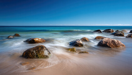A picturesque beach scene featuring gentle waves washing over rocks under a bright blue sky, showcasing natural coastal beauty.