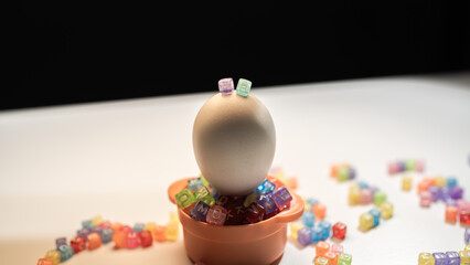 A delicate white egg sits in a small orange pot filled with colorful alphabet beads. It represents...