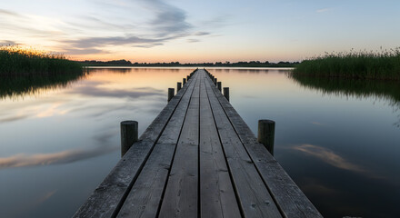 Obraz premium Peaceful wooden pier stretching into tranquil lake at sunset, reflecting the colorful sky and calm waters