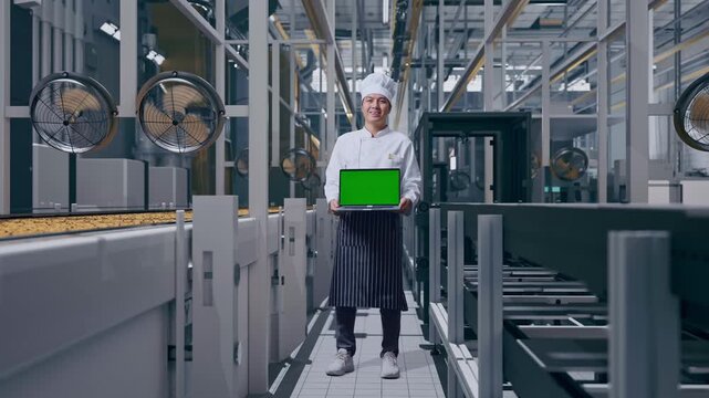 Full Body Of Asian Man Chef Smiling And Showing Green Screen Laptop To The Camera While Standing, Automated Cookie Production Line in Modern Factory