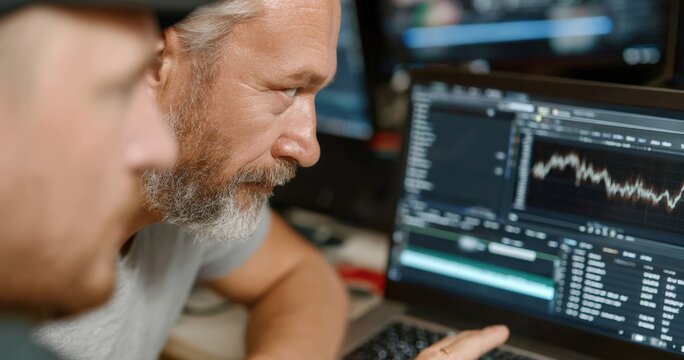 Two software developers concentrating while analyzing code and graph on computer screen, working together in office at night