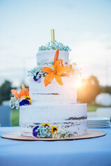 Three-tiered white wedding cake decorated with  orange, blue, yellow and white fresh flowers   on a wooden stand with fireworks on top. Side view