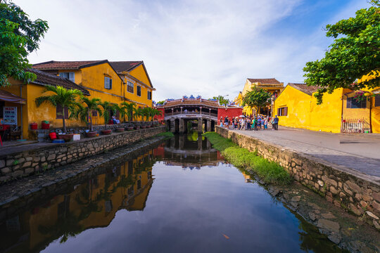 A scenic view of the iconic Chua Cau bridge reflecting on a small canal, surrounded by traditional yellow buildings and lush greenery on a peaceful day in Hoi An.
