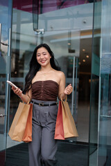 Happy customer exiting shopping mall holding shopping bags and smartphone