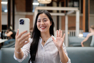 Young Asian businesswoman making a video call and waving at smartphone in the office