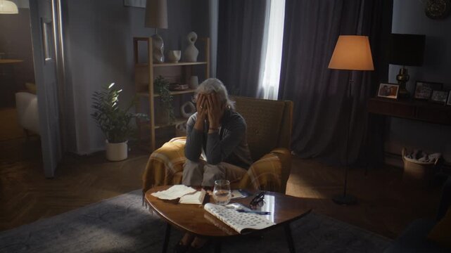 Desperate elderly woman hides face behind palms in armchair