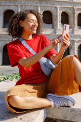 A cheerful young woman sits on a stone ledge, happily using her smartphone, enjoying the beautiful view of the ancient Colosseum in Rome, blending rich culture with modern technology