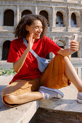 A joyful young woman in casual clothes sits on a stone ledge, waving while taking a selfie in front of the iconic Colosseum in Rome, capturing a wonderful moment of her travel adventure