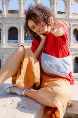 A joyful young woman poses playfully against the backdrop of the iconic Colosseum, proudly showcasing her stylish outfit and vibrant personality while capturing the moment in a delightful selfie