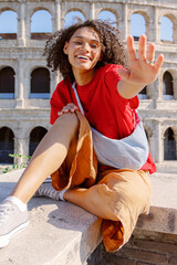 A joyful young woman waves at the camera while sitting on a stone ledge, with the Colosseum in the backdrop, showcasing her vibrant personality during her sightseeing adventure in Rome