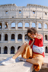 A joyful young woman in adventurous attire sits by the historic Coliseum, making peace signs...
