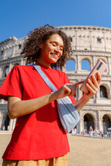 A cheerful young woman in a bright red shirt happily uses her smartphone in front of the stunning Colosseum in Rome, capturing the joy of her travels and youthful spirit in this beautiful spot