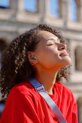 A joyful woman with beautiful curly hair, dressed in a bright red shirt, basks in the warm sunlight at a breathtaking landmark, radiating warmth and serenity during this truly peaceful moment