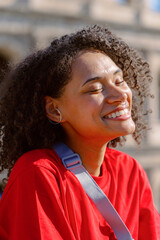 On a sunny day, a vibrant young woman with curly hair enjoys the outdoors in a bright red shirt and modern earbuds, radiating happiness and a carefree vibe that captivates those around her