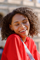 A cheerful young woman with curly hair in a stylish red shirt radiates joy outdoors, positively affecting others and creating a warm, inviting atmosphere with her infectious happiness and energy