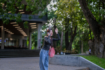 Young woman talking on phone and waving in park near university building