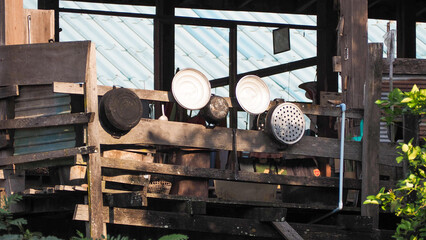 weathered, wooden-slatted outdoor structure, possibly an informal kitchen or utility area in a rural setting, featuring household items like pots, pans, and showerheads (used as strainers) hung 
