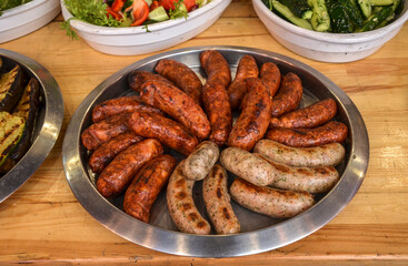 Silver platter of assorted grilled sausages, some browned and some lighter, is on wooden table. Other dishes, including salads with tomatoes, cucumbers, and other vegetables, are in the background
