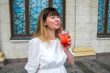 Young woman sips a cold fresh red lemonade with ice outdoors, showcasing relaxation and enjoyment...