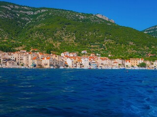 Fototapeta premium Komiža harbor on Vis Island, Dalmatia, Croatia—stone houses with terracotta roofs line the Adriatic, backed by lush green hills and clear blue water under a bright summer sky.