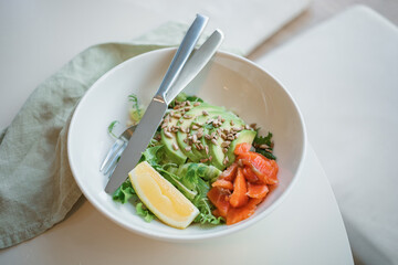 Close-up of  a bowl with green salad with smoked salmon, lettuce and avocado, garnished with seeds and lemon.