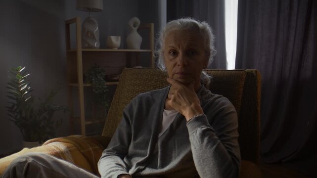 Pensive senior woman sits thoughtfully in armchair in living room