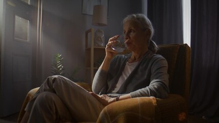 Elderly woman drinks water from glass sitting in vintage armchair - Powered by Adobe