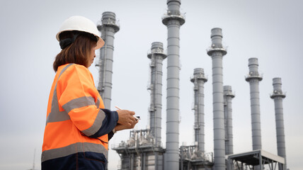A worker equipped with a hard hat and high-visibility jacket stands near a power plant, observing tall smokestacks and recording observations while on duty © Happy Photo