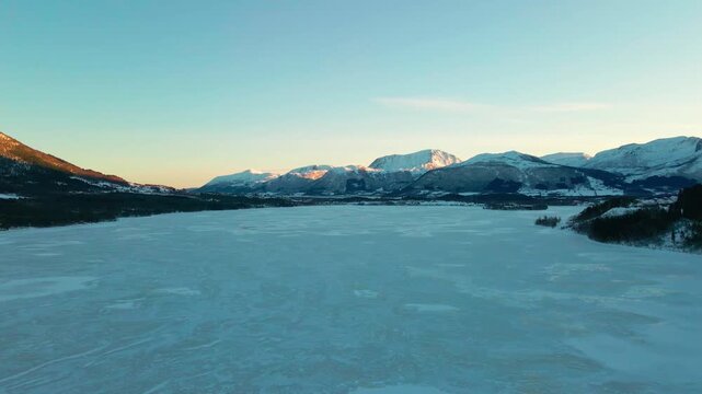 An aerial view captures the serene beauty of a vast, frozen fjord or lake surrounded by snow capped mountains under a vibrant sunrise sky, evoking the tranquil winter scenery of a remote Norwegian fis