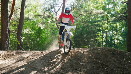 A motorcyclist in a red shirt rides a dirt bike through a sunny forest