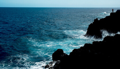 Person standing on rock cliffs above crashing waves below